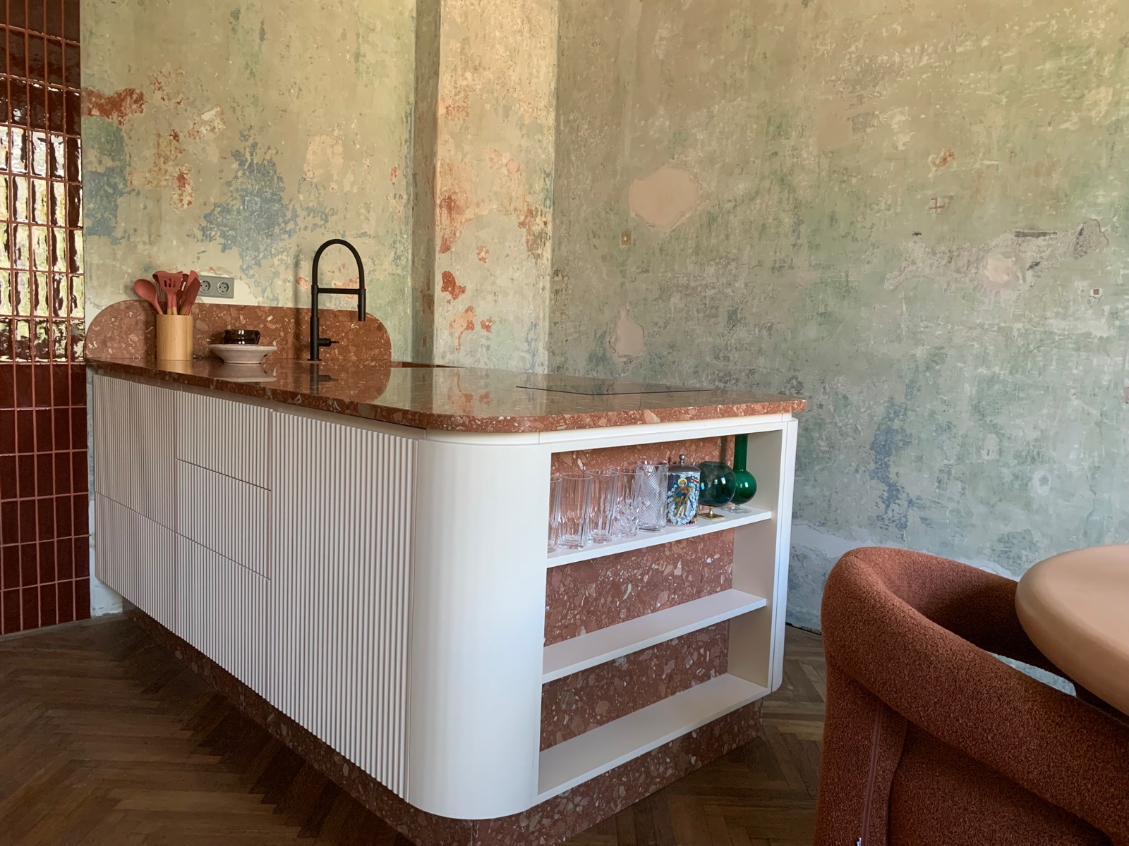 Moderne Theke mit gerundeten Ecken, weiße Wellpappe-Front, Terrazzo-Arbeitsplatte in Rot und Grau. Im Hintergrund abblätternde Wand in Grün, Ocker und Rostrot. Links ein rotes Fachwerk-Fenster. Auf der Theke ein Terrakotta-Gefäß, eine weiße Schale, ein schwarzer Wasserhahn. Rechts im Bild ein Sessel in Braun-Rot.